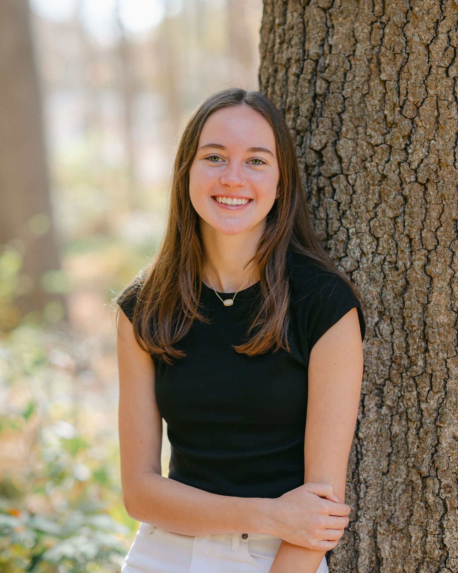 Portrait of person in black top leaning against large tree trunk in wooded setting.