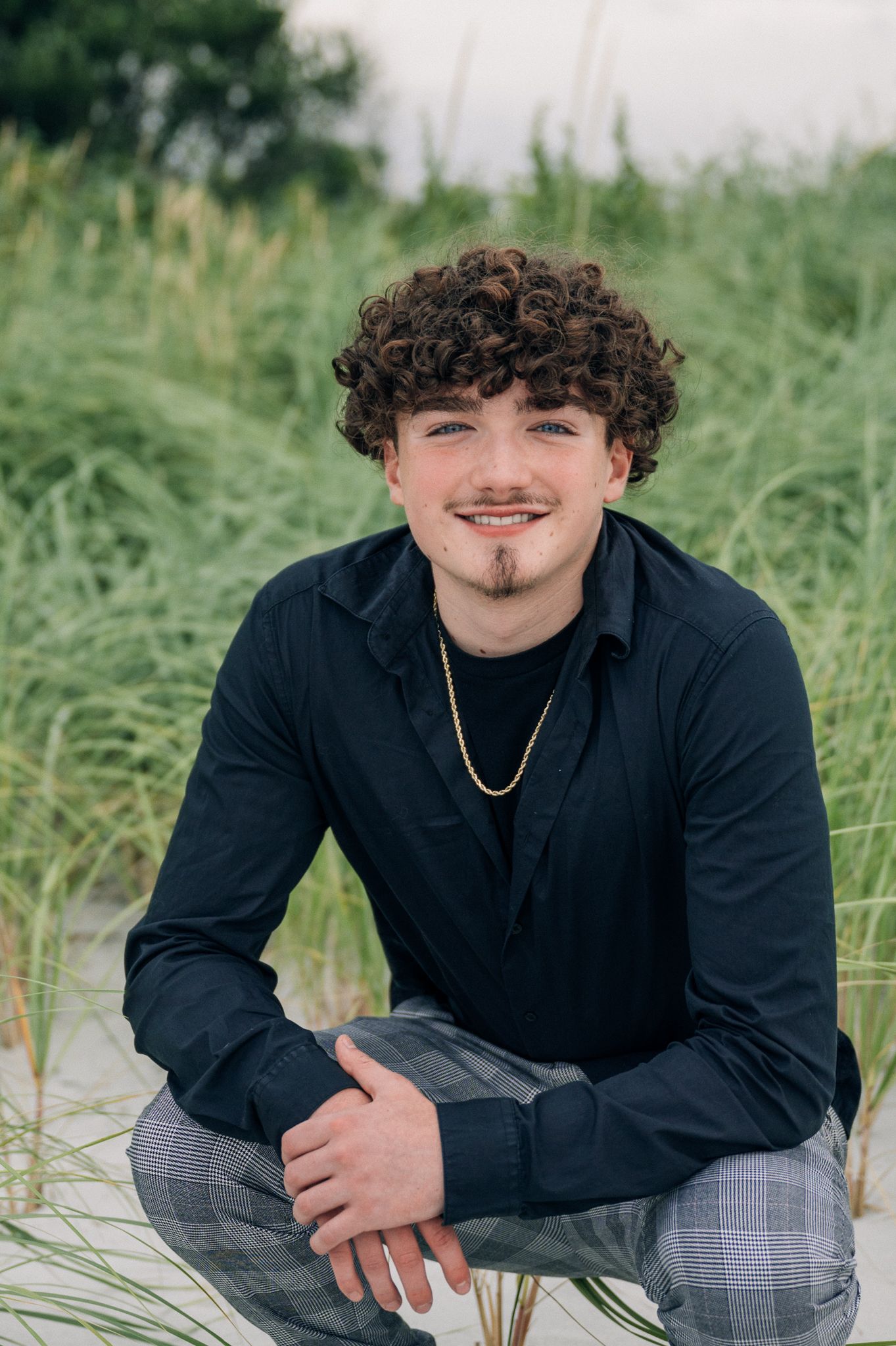 Casual portrait of a person in dark clothing and plaid pants seated in beach grass.