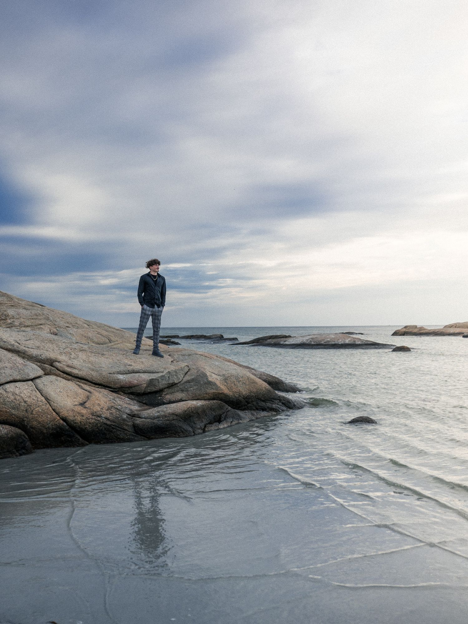 Silhouetted figure stands on rocky coastal outcrop overlooking ocean under dramatic cloudy sky.