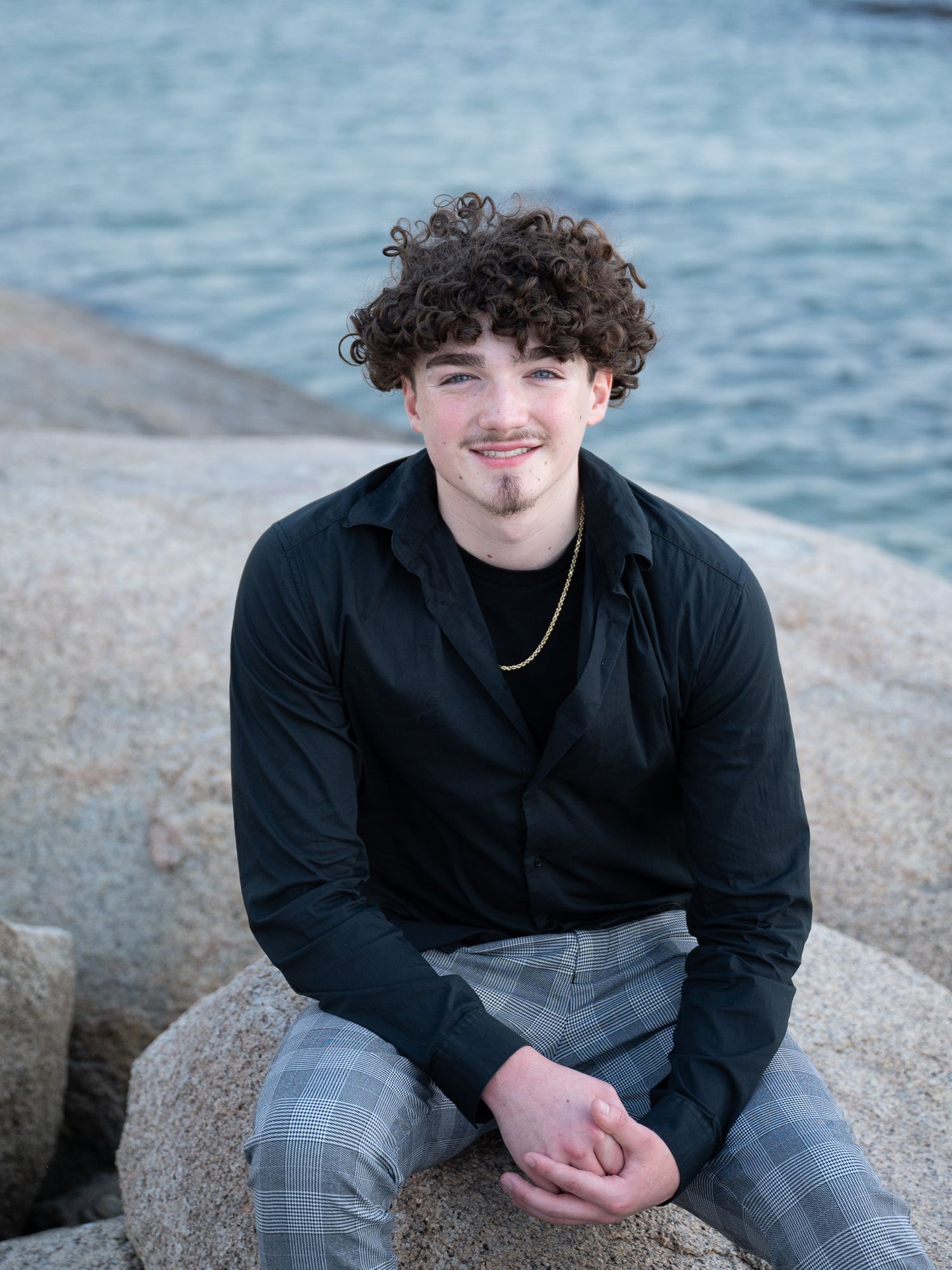 Casual seaside portrait with person seated on coastal rocks wearing dark shirt and plaid pants.