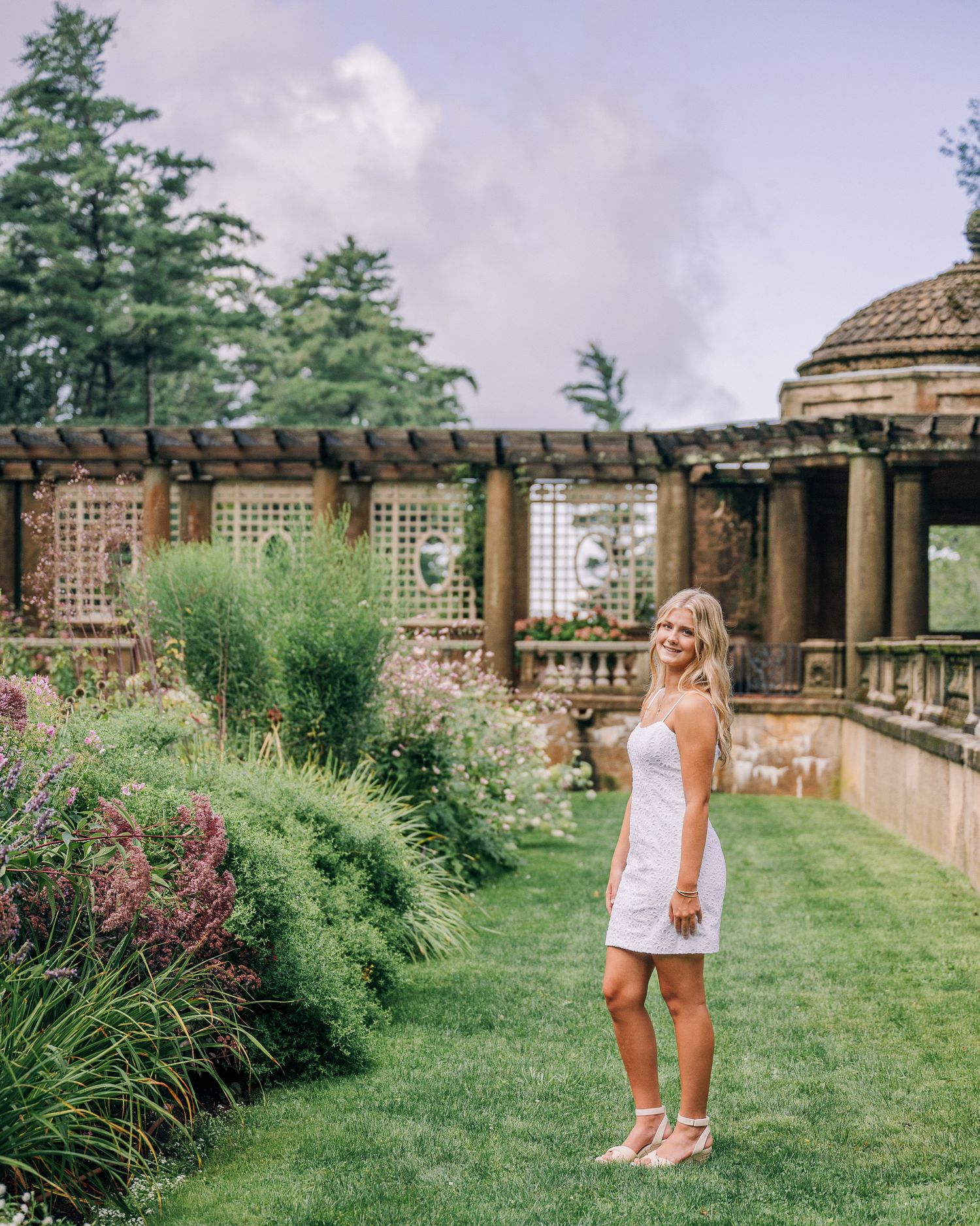 A white dress adorned figure stands on manicured lawn near ornate garden pergola with lattice screens and stone columns.