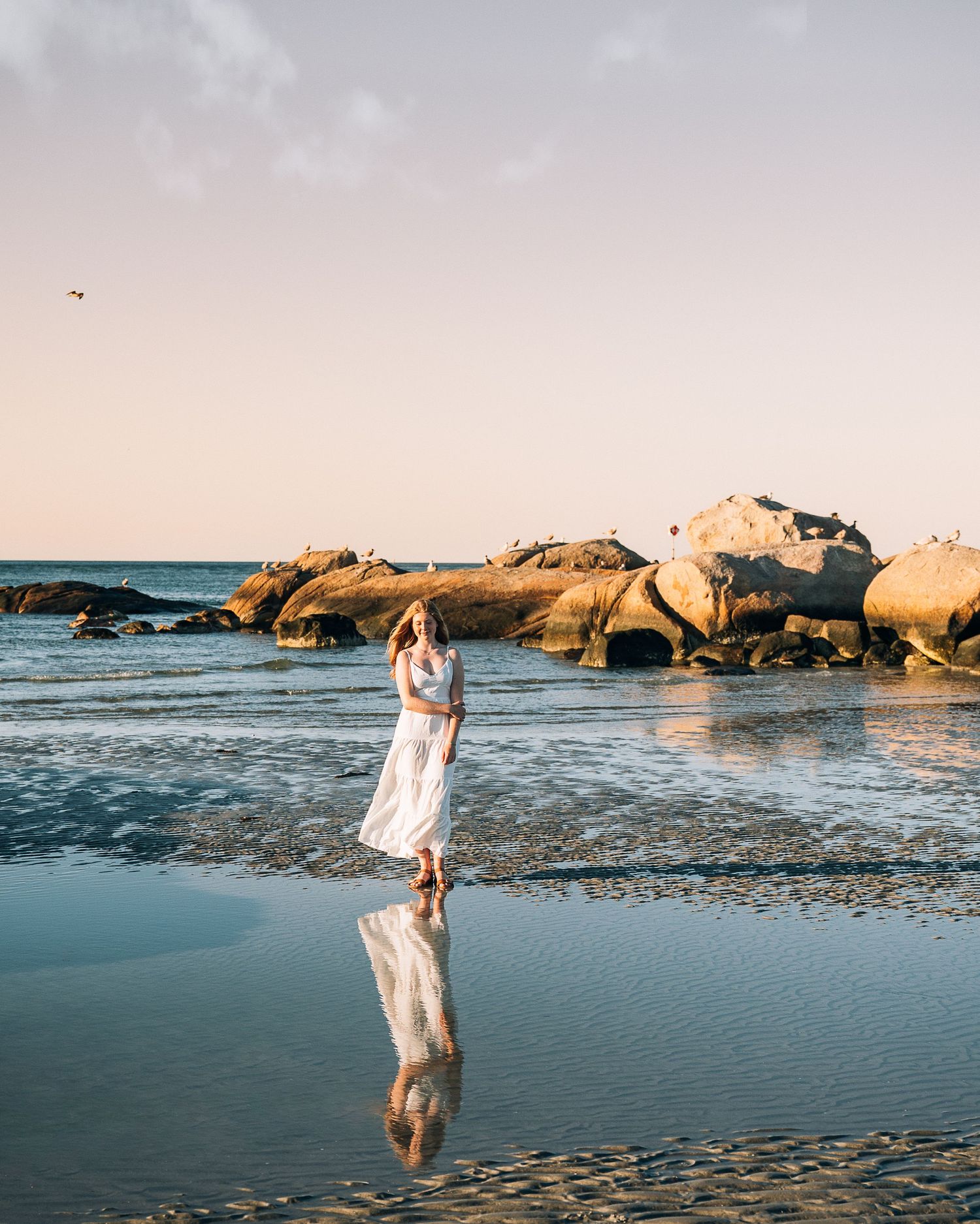 Woman in white maxi dress walking on reflective wet sand at sunset with rocky coastline.