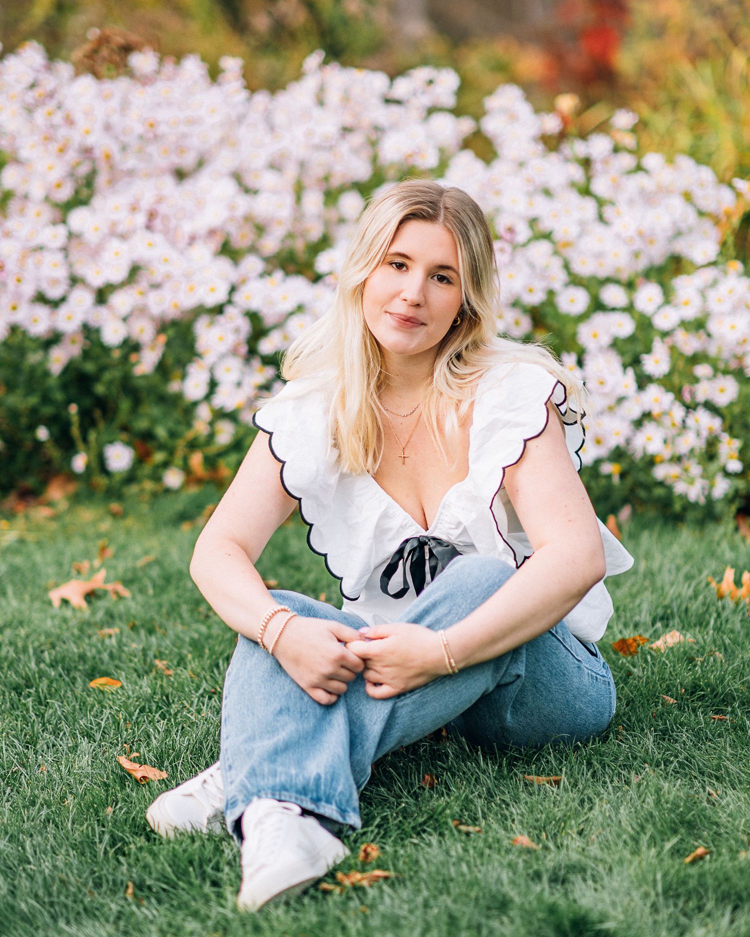 Casual portrait in ruffled top and jeans sitting on grass with flowers in background.
