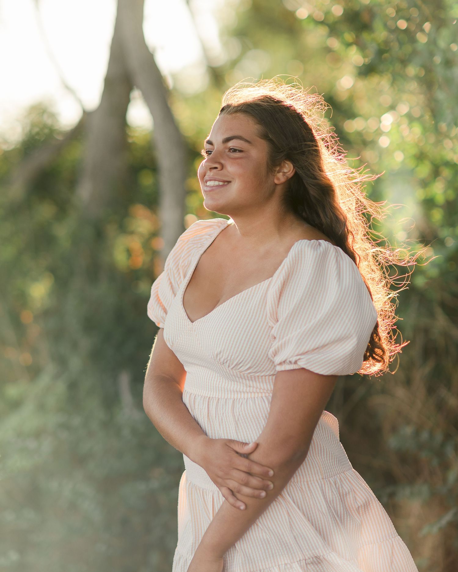 Person in white dress poses joyfully in garden setting with backlit golden sunlight.