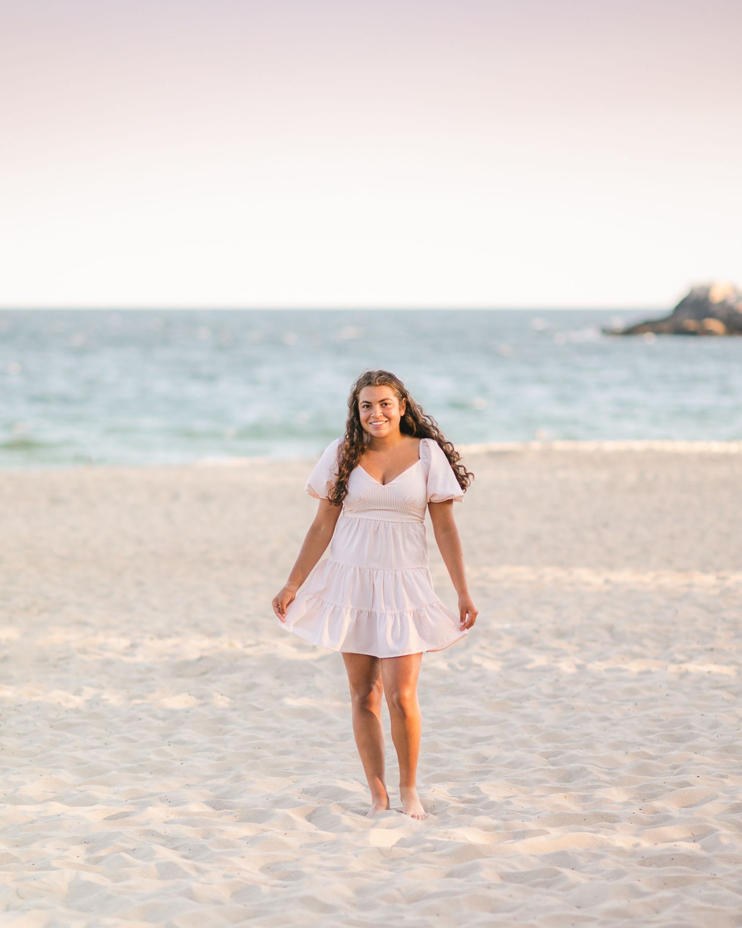 A person in a white summer dress walks barefoot along a sandy beach at sunset.