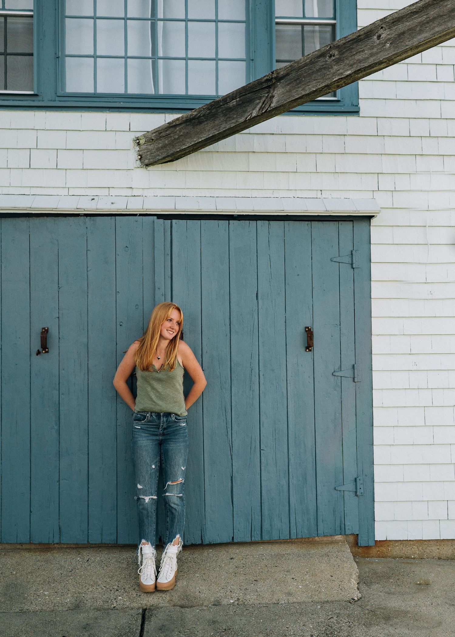 A person in tank top and ripped jeans poses against a blue barn door and white building.