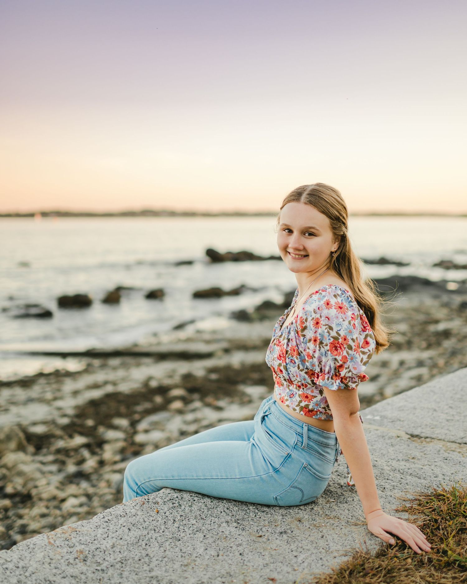 Person in floral top and jeans relaxes on coastal rocks at sunset.