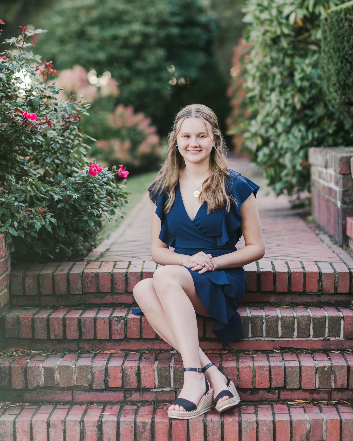 Person in navy blue dress sits elegantly on brick steps surrounded by flowering garden bushes.