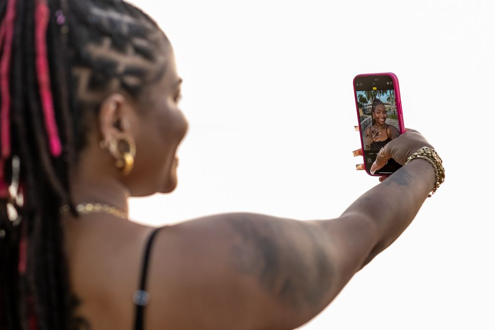 Person taking a selfie with a red phone case against a blurred outdoor background.