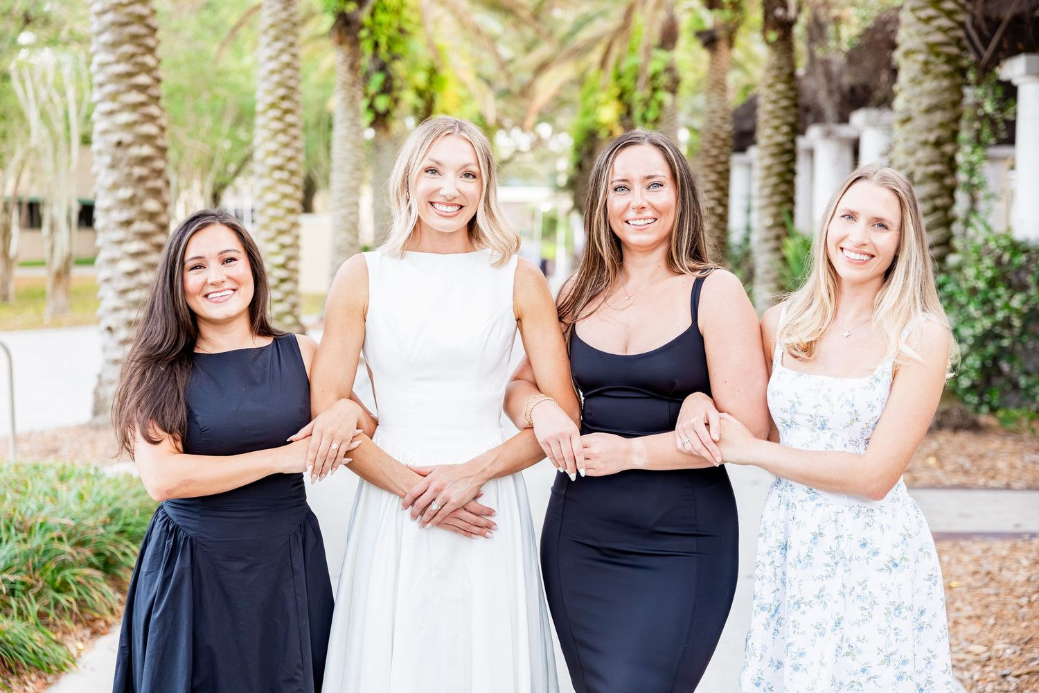 Four smiling women in elegant dresses posing together under palm trees outdoors.