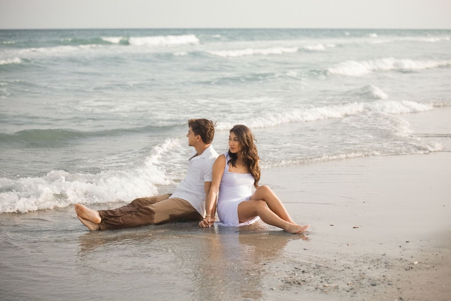 Relaxed couple sitting at water's edge on beach in casual attire.