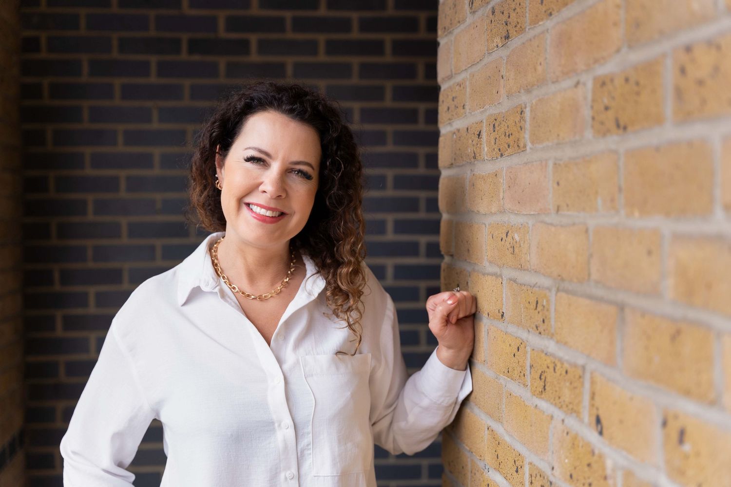 Woman wearing white button-up shirt stands against a brick wall smiling brightly and confidently.