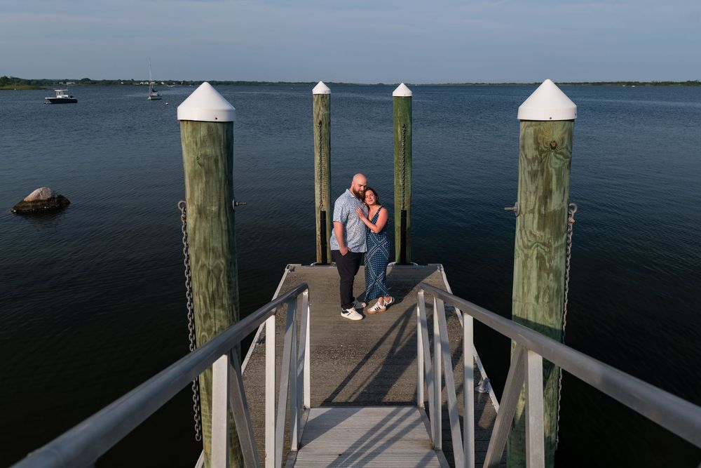 A couple stands together on a wooden dock extending into calm waters at dusk, surrounded by mooring posts.