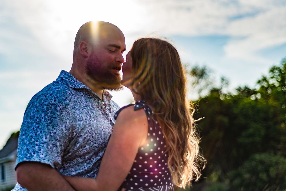 A couple shares an intimate moment at sunset with trees and a house in the background.