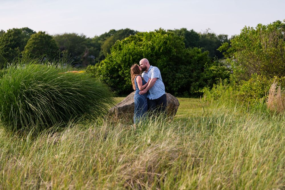 A romantic couple sits together in a scenic grassy field during golden hour surrounded by tall grass and sunlit nature.