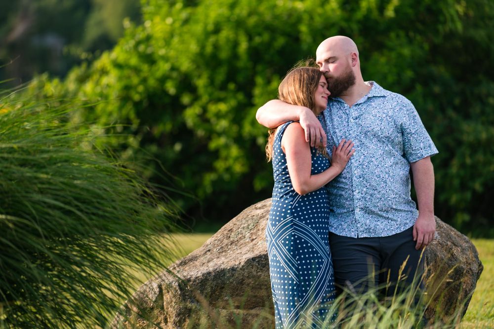 A romantic couple in blue outfits embraces while sitting on a grassy hill during a sunny outdoor photoshoot.