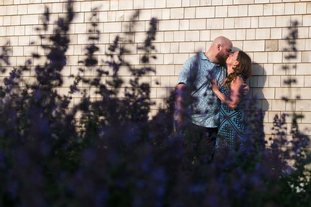 Person wearing denim jacket stands against brick wall with dappled sunlight creating shadow pattern effect.