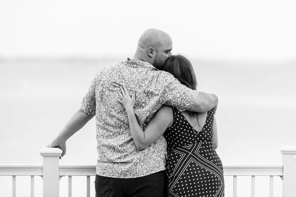 Two people in black and polka dot dresses embrace on an outdoor patio near a white railing with ocean views.