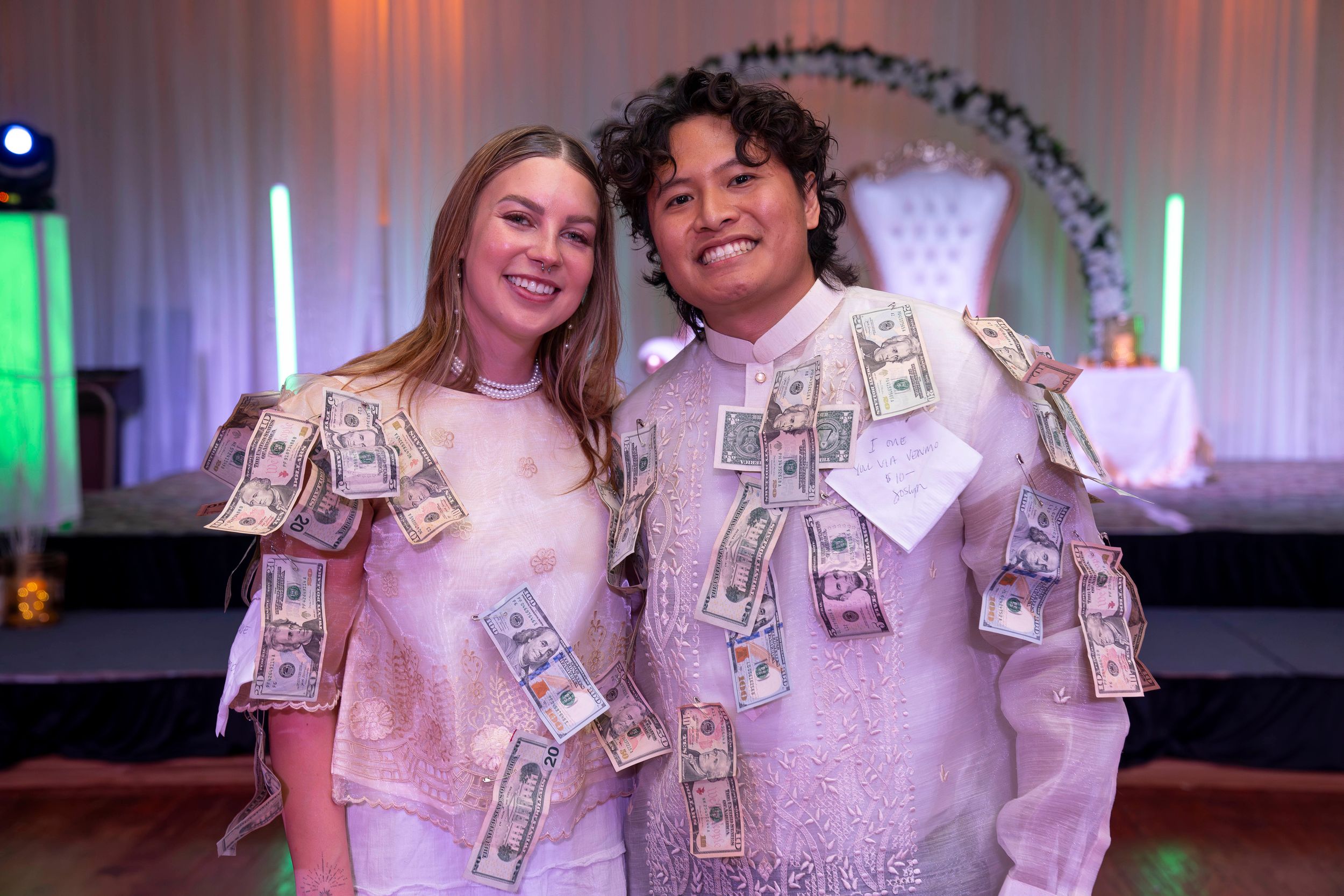 Two people wearing money pinned to their outfits pose together at a celebration event with decorative lighting.