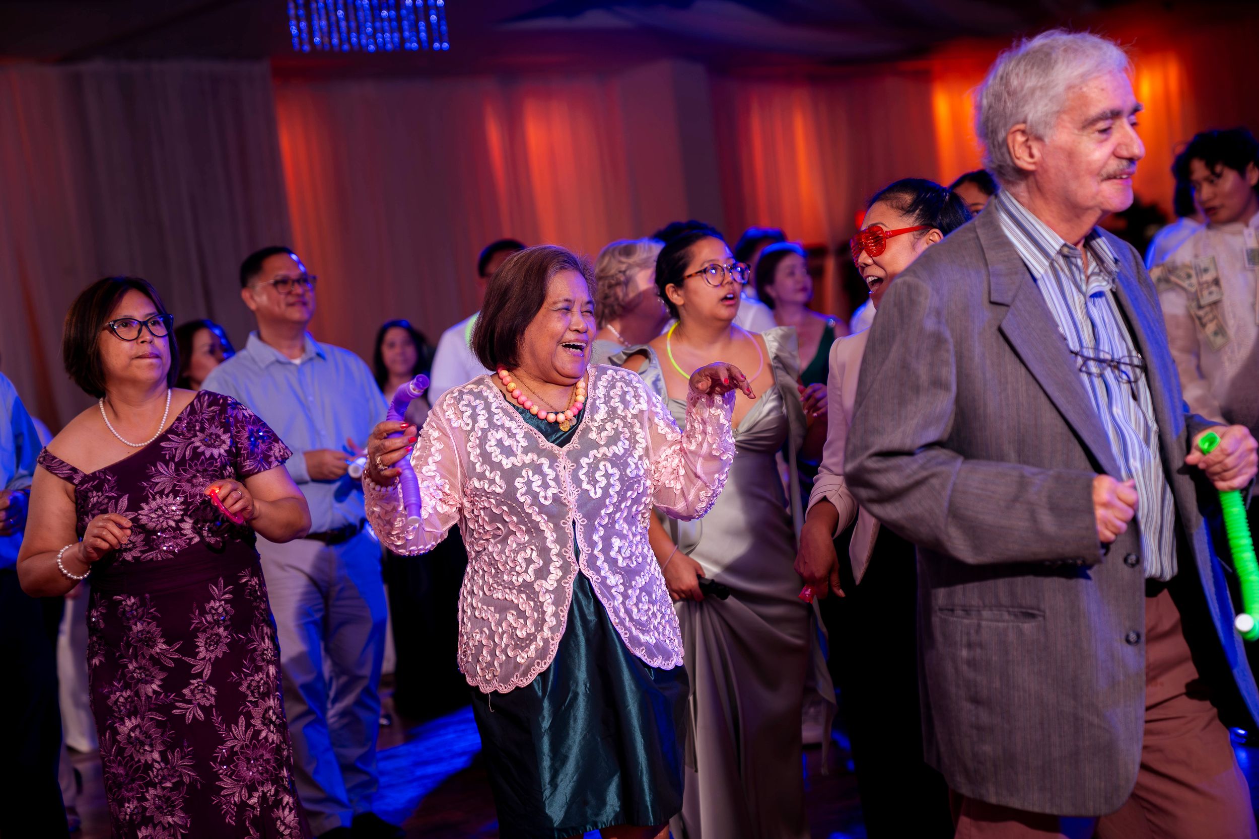 Wedding guests dancing and celebrating on a dance floor illuminated by red and blue lighting.