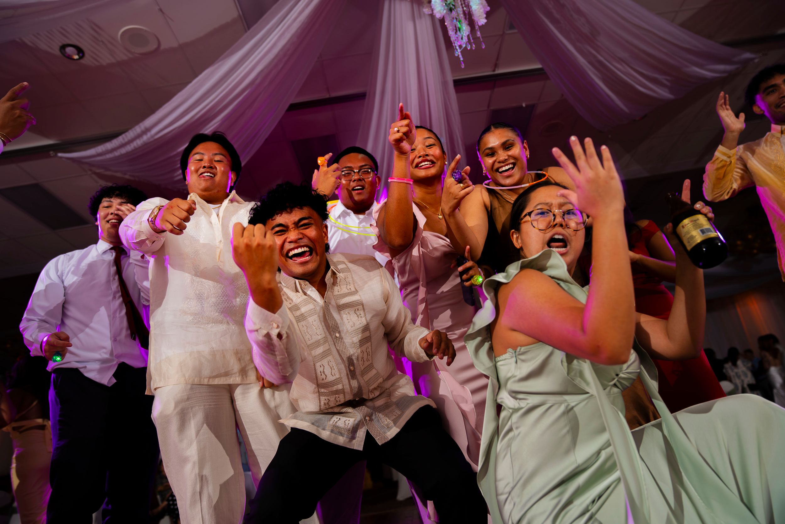 Wedding party dancing energetically and joyfully at a reception under draped white fabric ceiling decorations.