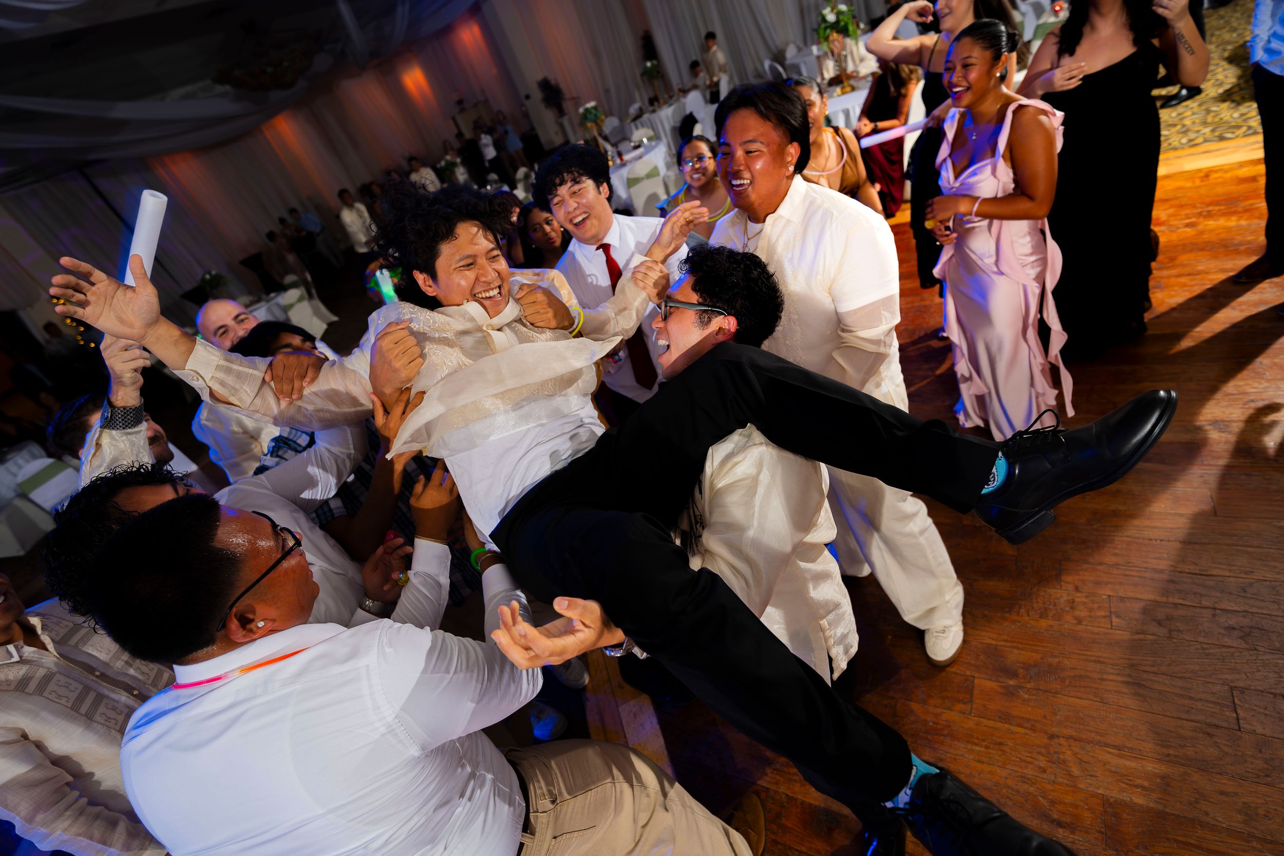 Wedding reception guests show joyful dance moves on a wooden dance floor in dimly lit indoor venue.
