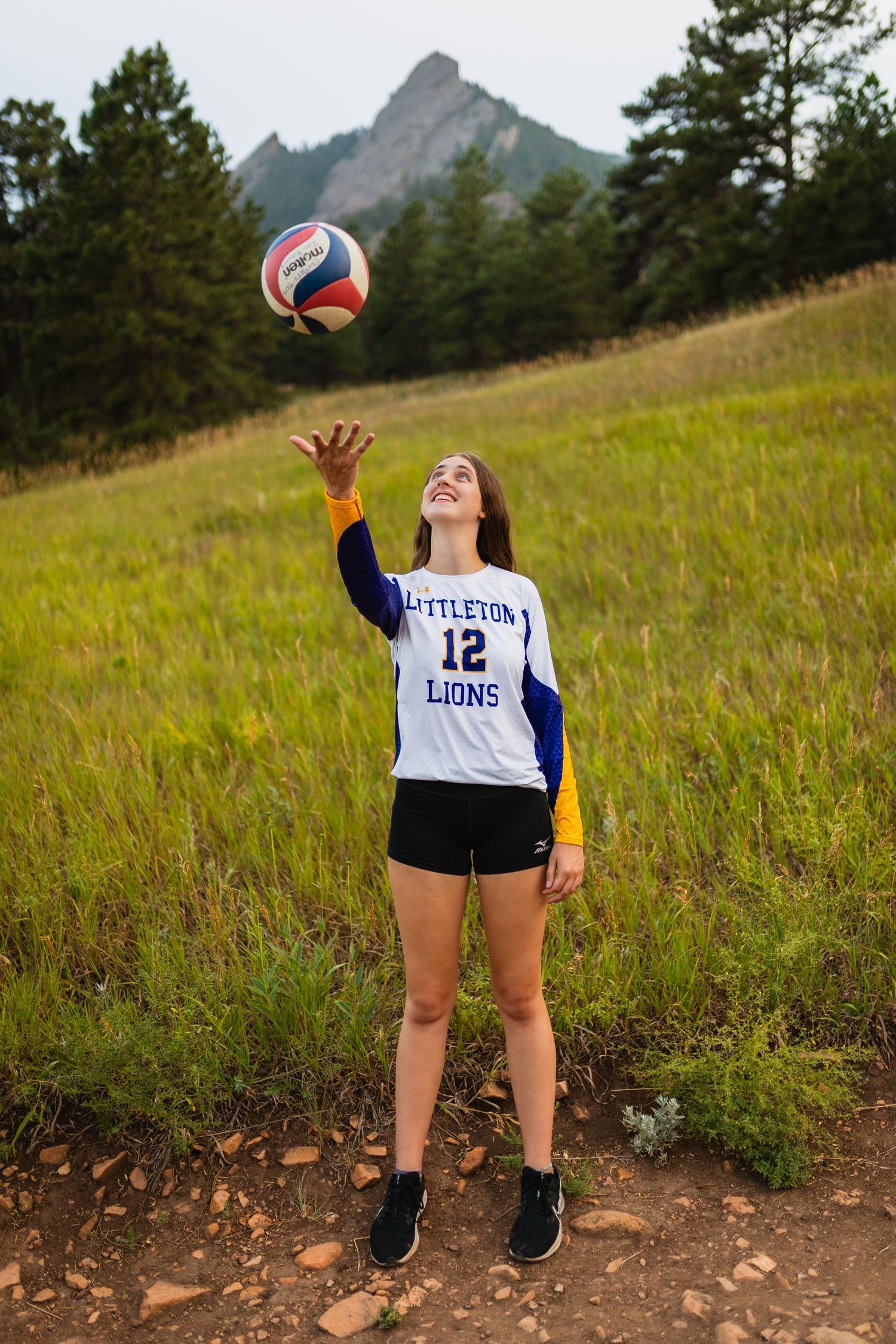 A volleyball player practices serves on a scenic mountain trail surrounded by tall grass and evergreen trees.