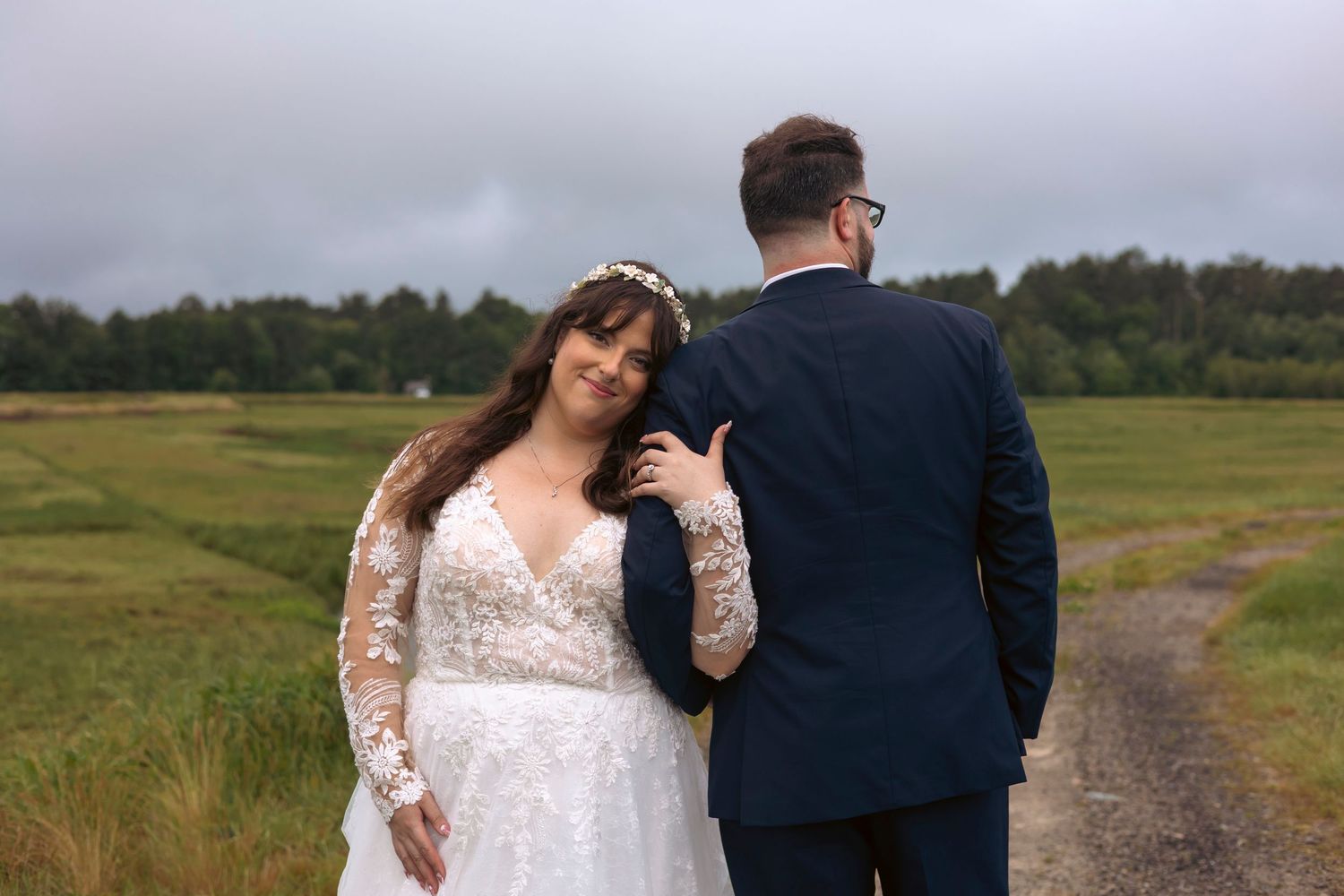 A couple in wedding attire stands together on a country path surrounded by scenic grasslands and trees.