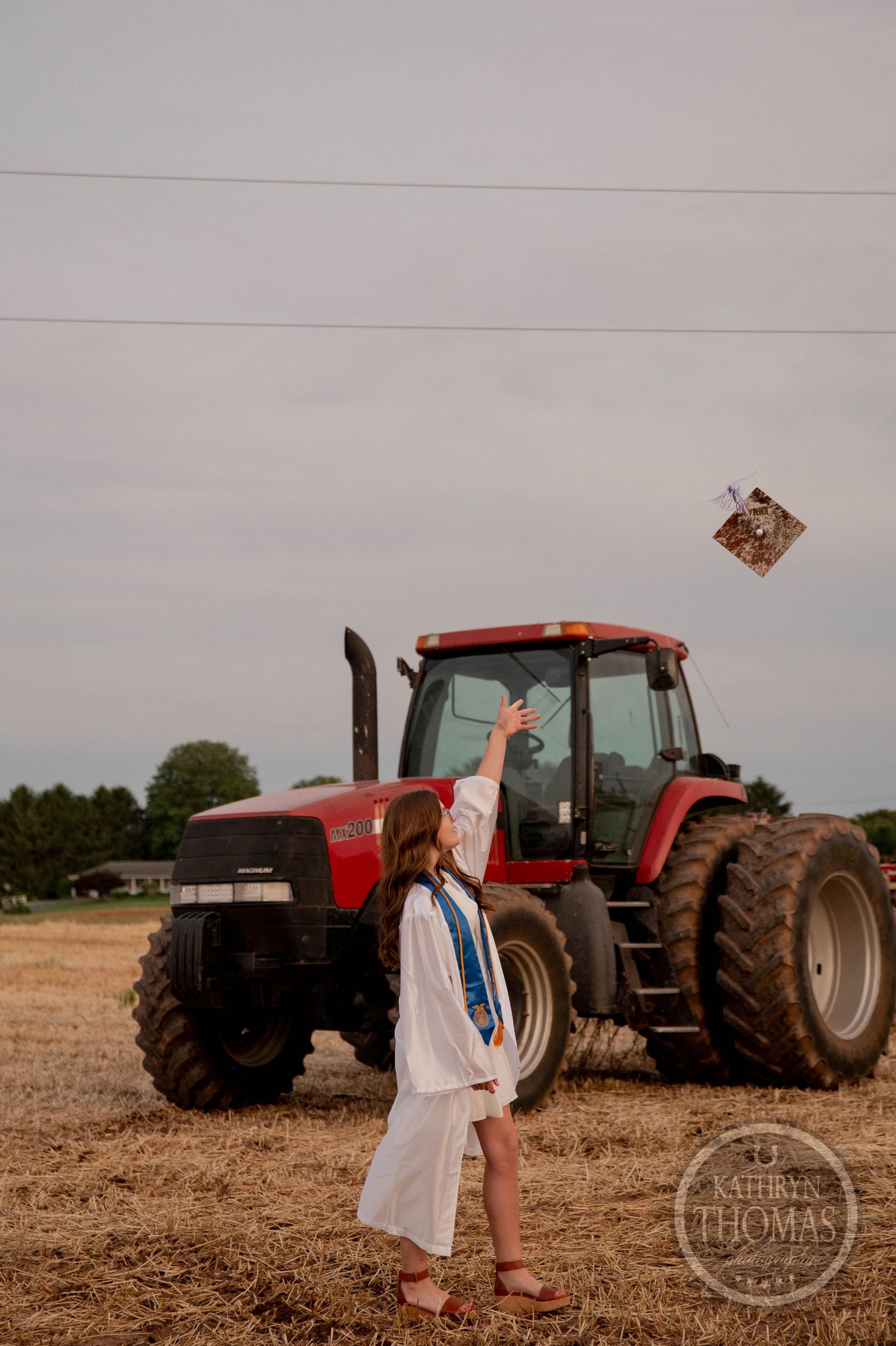 Red tractor parked in a harvested agricultural field at dusk with figures walking nearby against a cloudy sky.