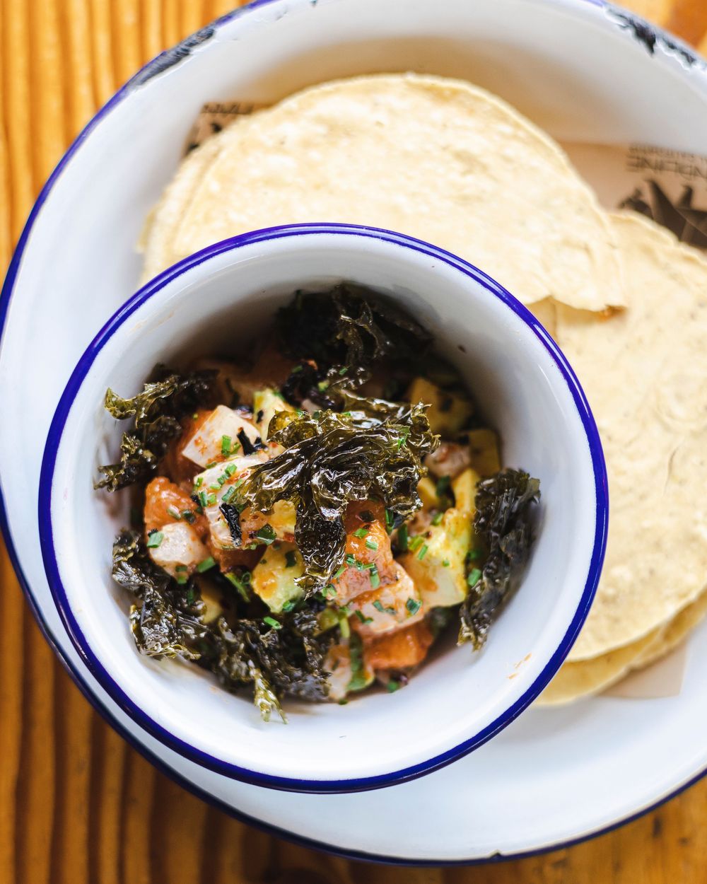 White enamel plates with blue rims containing sautéed shrimp and greens served alongside tortillas.