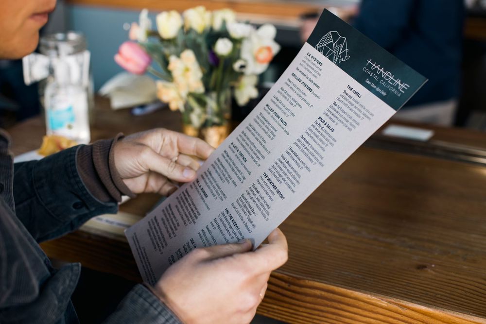 Someone reading a restaurant menu at a wooden table while making dining selections.