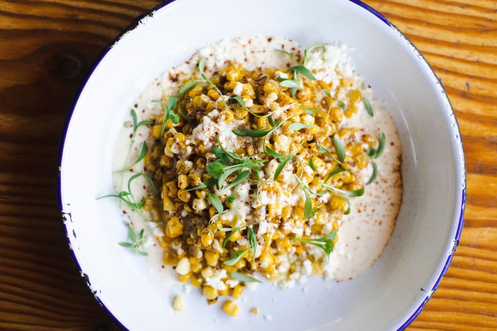 A white bowl of rice topped with corn kernels, fresh herbs and seasonings served on a wooden table.