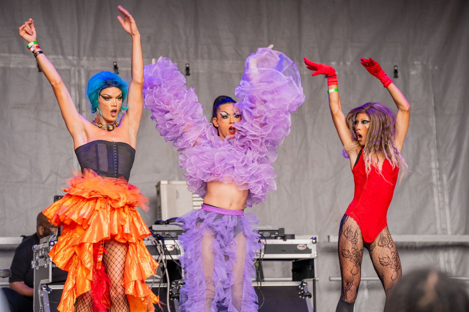 World Pride blue haired drag queen in an orange skirt stands with her arms over her head next to a queen in purple and another one in red