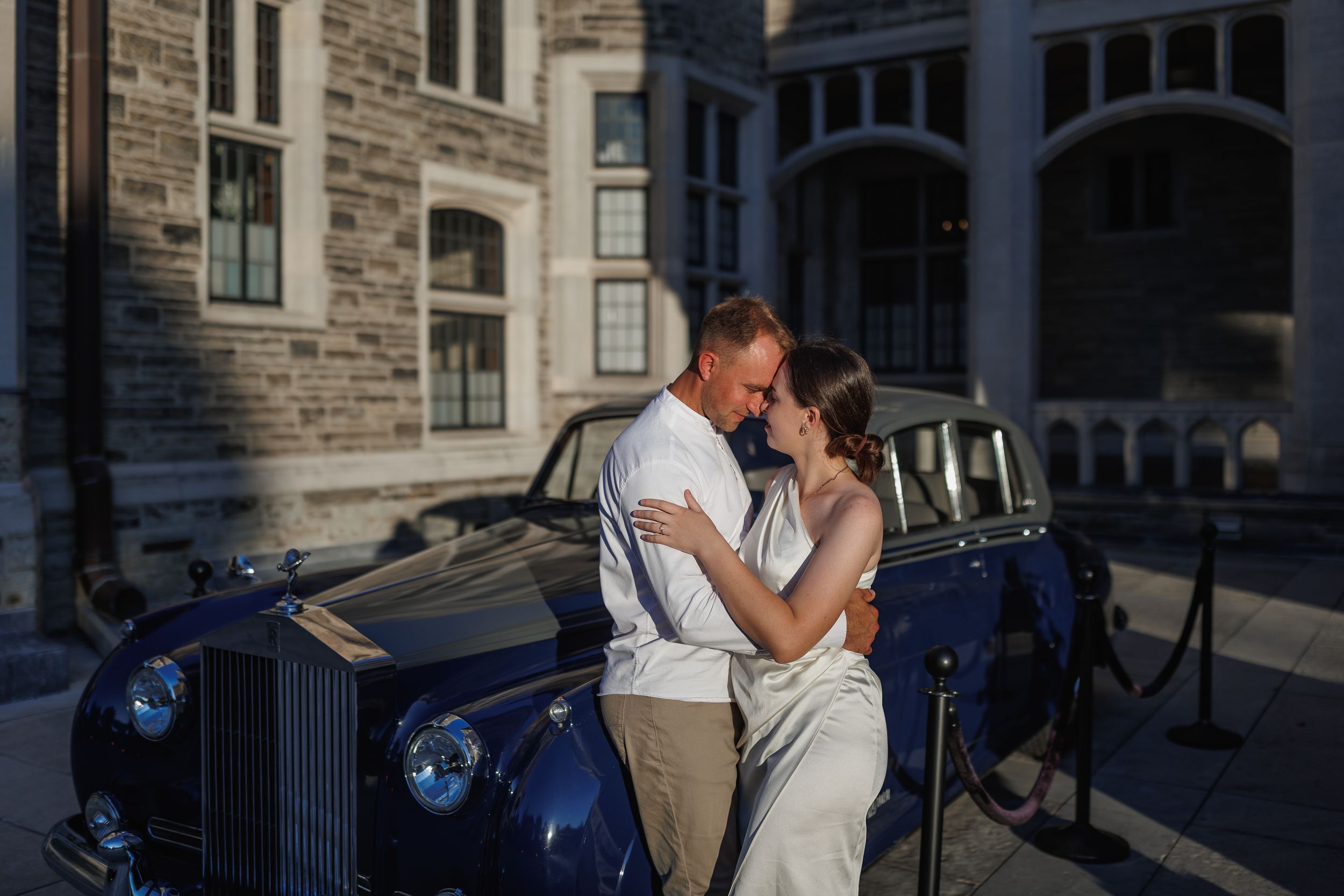 Couple embracing romantically in front of a vintage blue Rolls-Royce outside a stone building at golden hour.