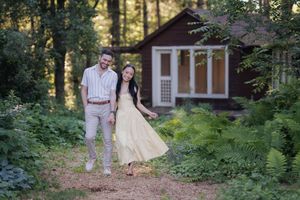 Couple smiling and walking hand-in-hand on a forest path near a rustic red cabin surrounded by lush greenery.