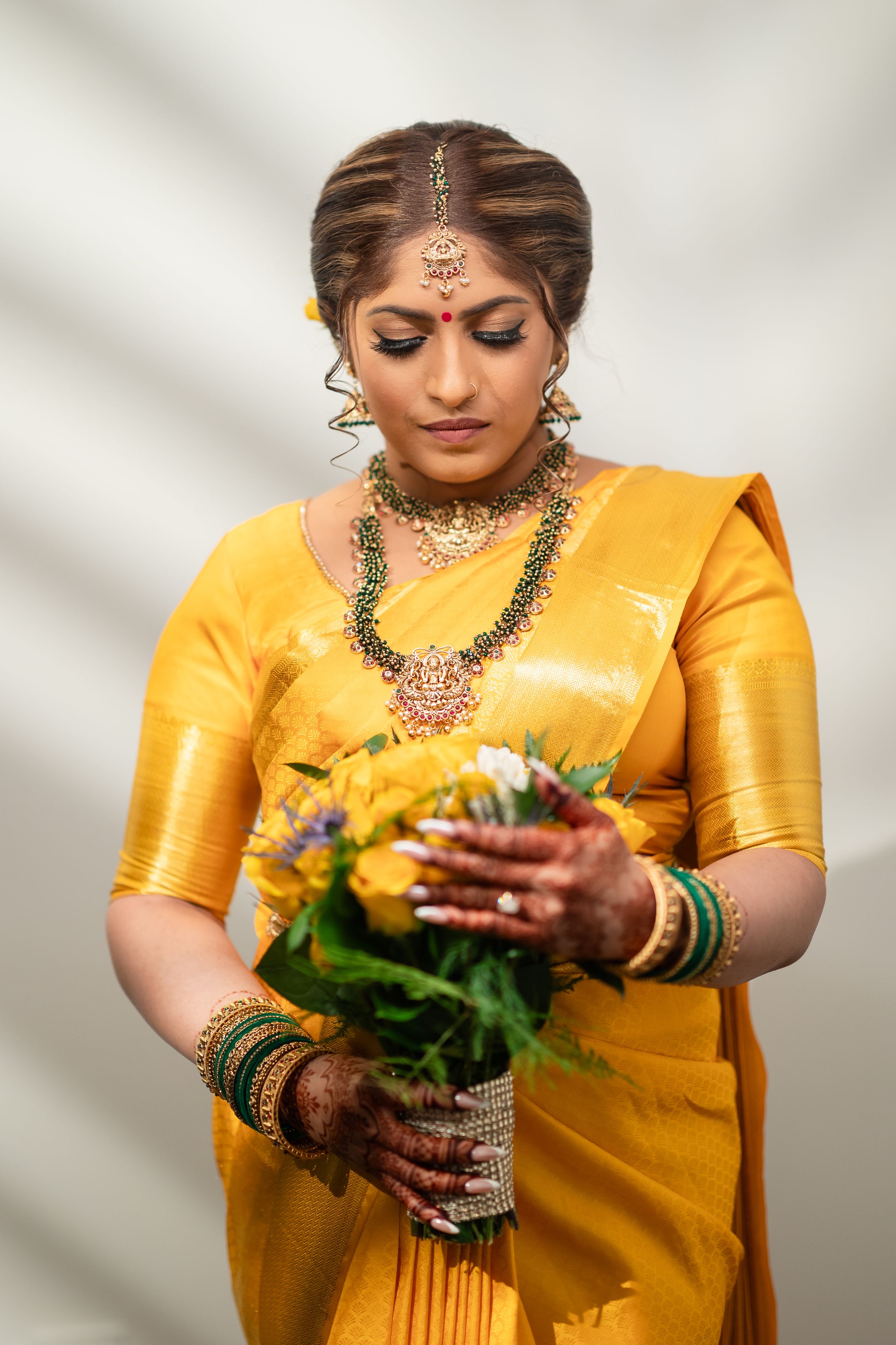 Traditional bride in yellow silk sari and ornate jewelry holds a bouquet against a light background.