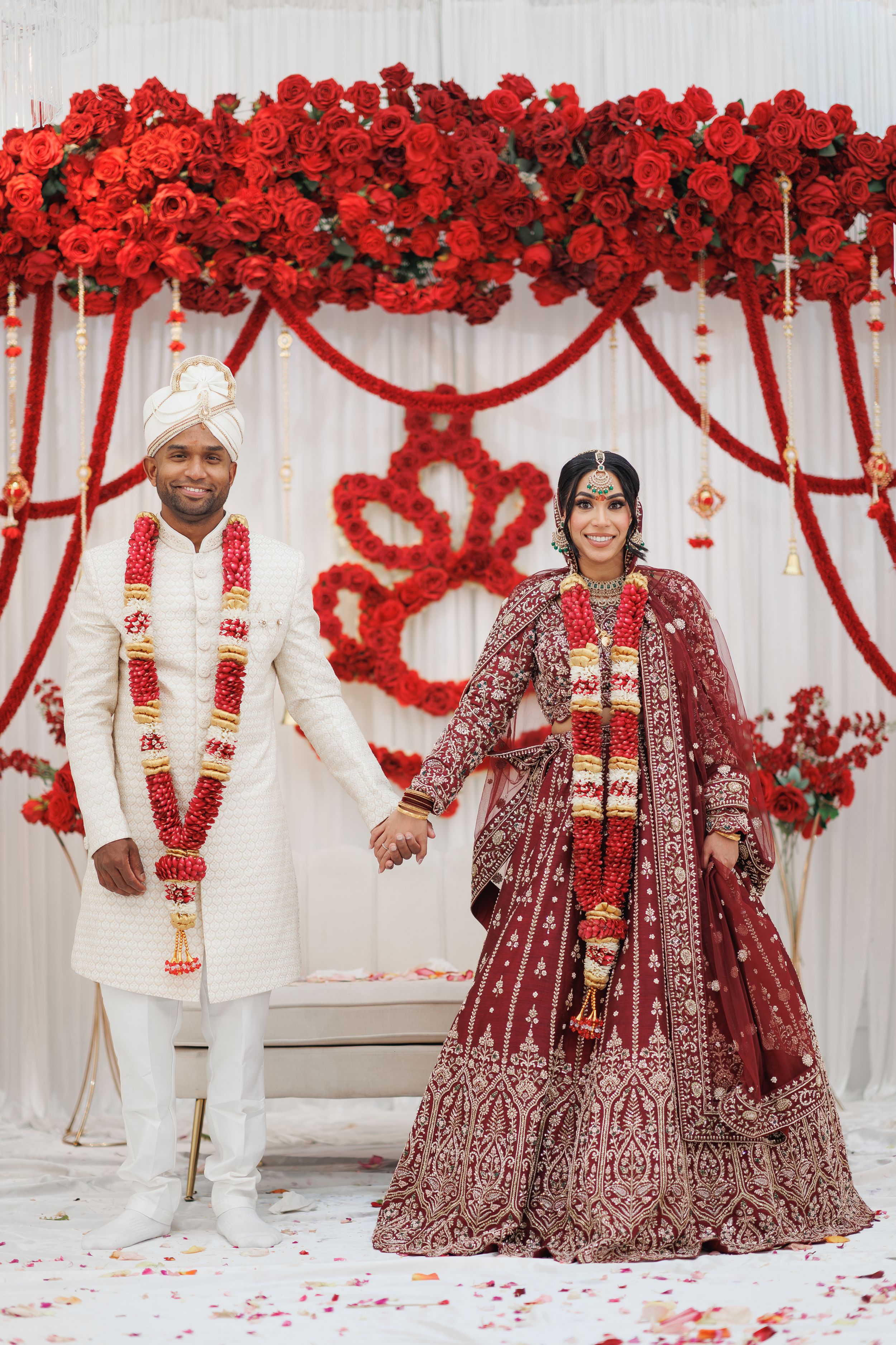 Indian bride and groom in traditional wedding attire pose before a stunning red rose floral arch backdrop.
