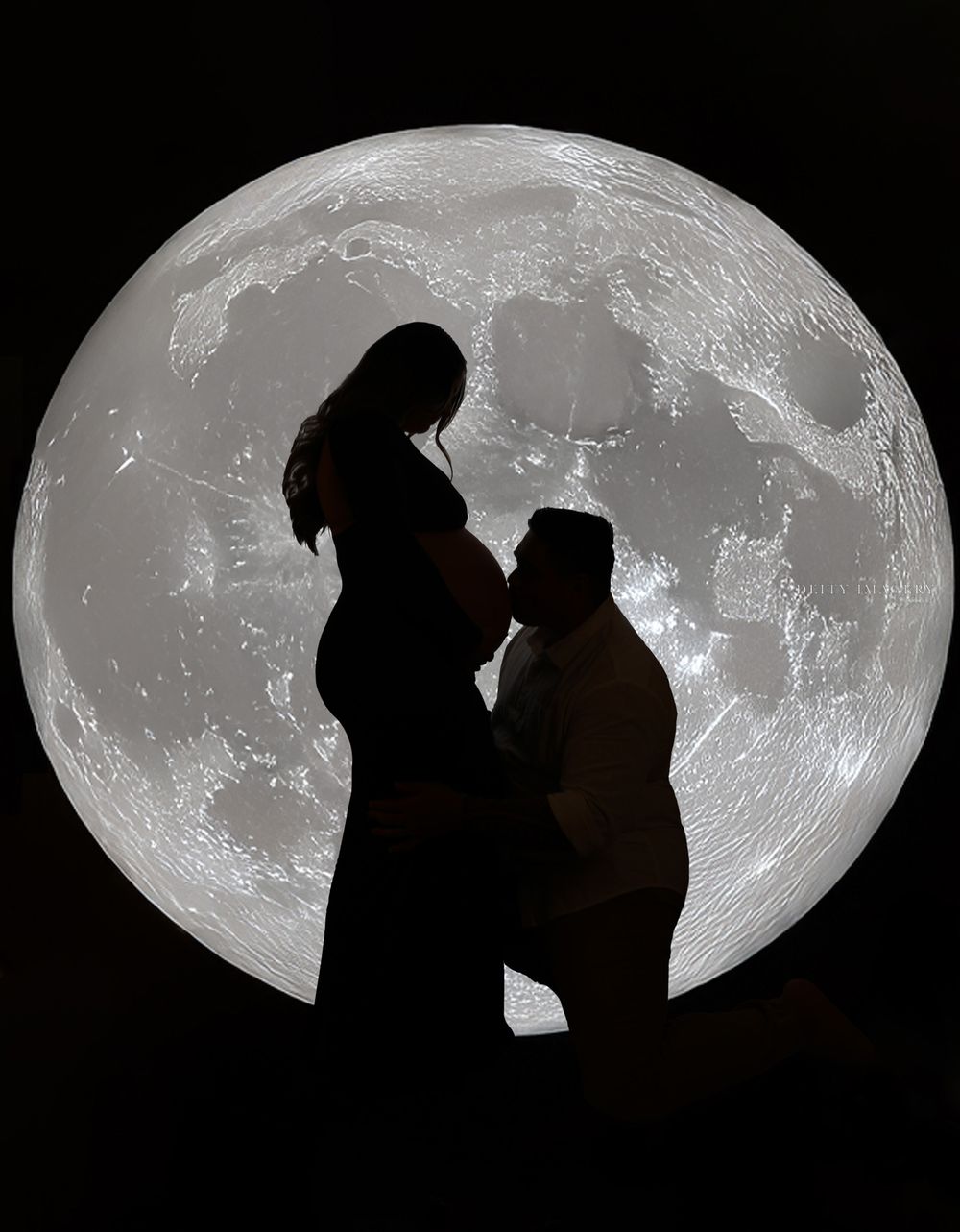 Silhouetted couple poses against large moon backdrop in artistic maternity series.