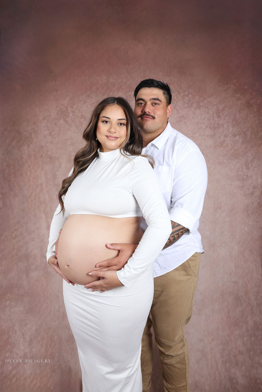 Professional maternity photoshoot showing a couple in white and khaki against a brown backdrop.