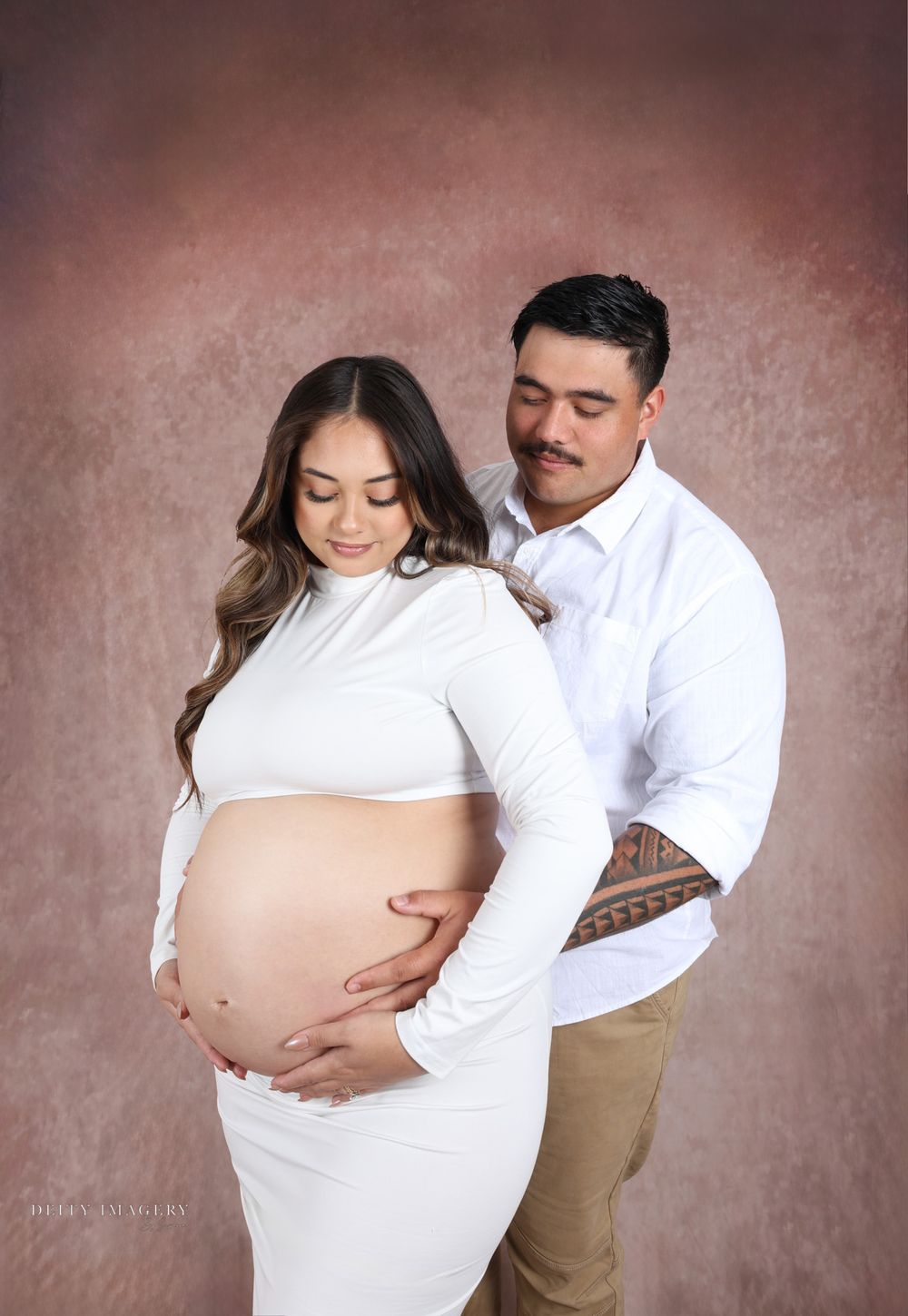 Intimate maternity portrait with couple in white and khaki clothing against textured brown background.