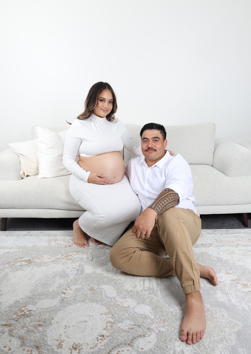 Modern maternity photo of a couple relaxing on white couch in minimalist setting.