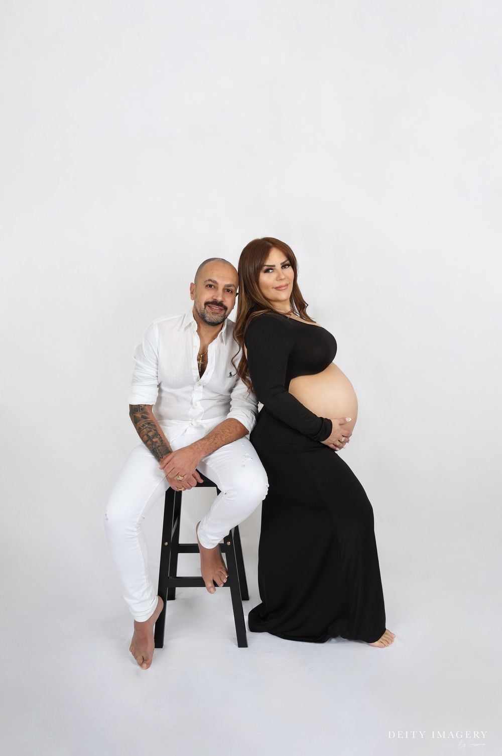 A pregnancy maternity photo shoot features a couple posing together in white and black outfits against a light backdrop.