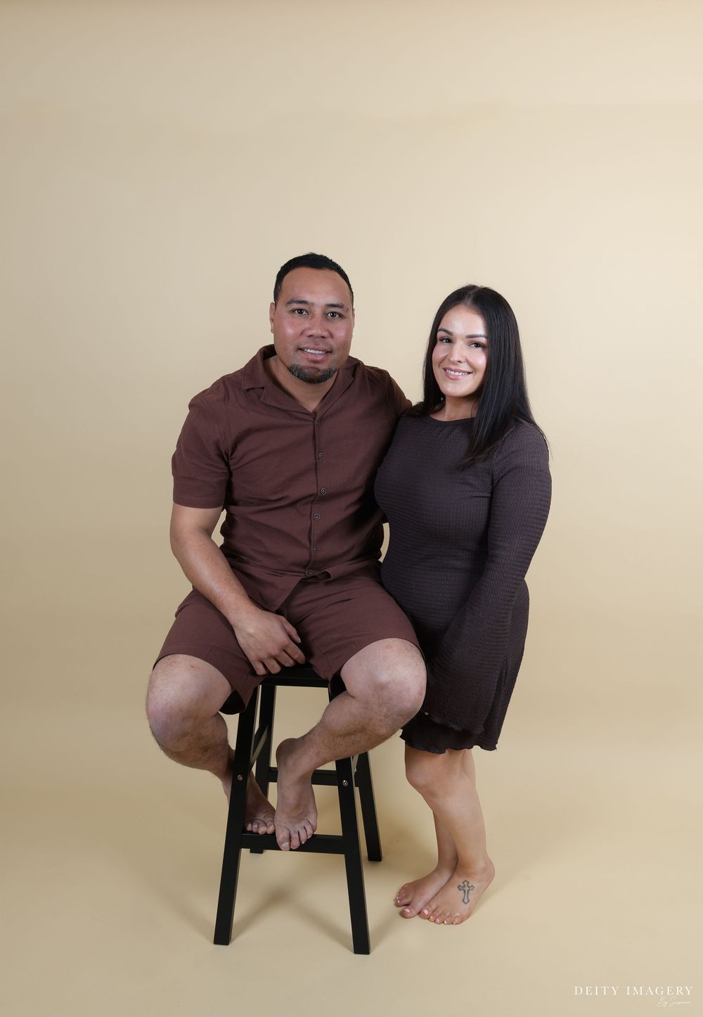 A couple poses together against a beige backdrop, with one person seated on a black stool and both barefoot.