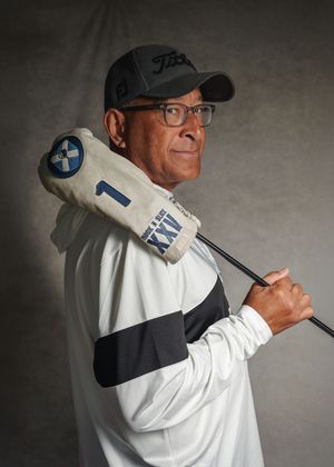A golfer in glasses wearing a black cap and white jacket poses with a golf club over their shoulder against a gray backdrop.