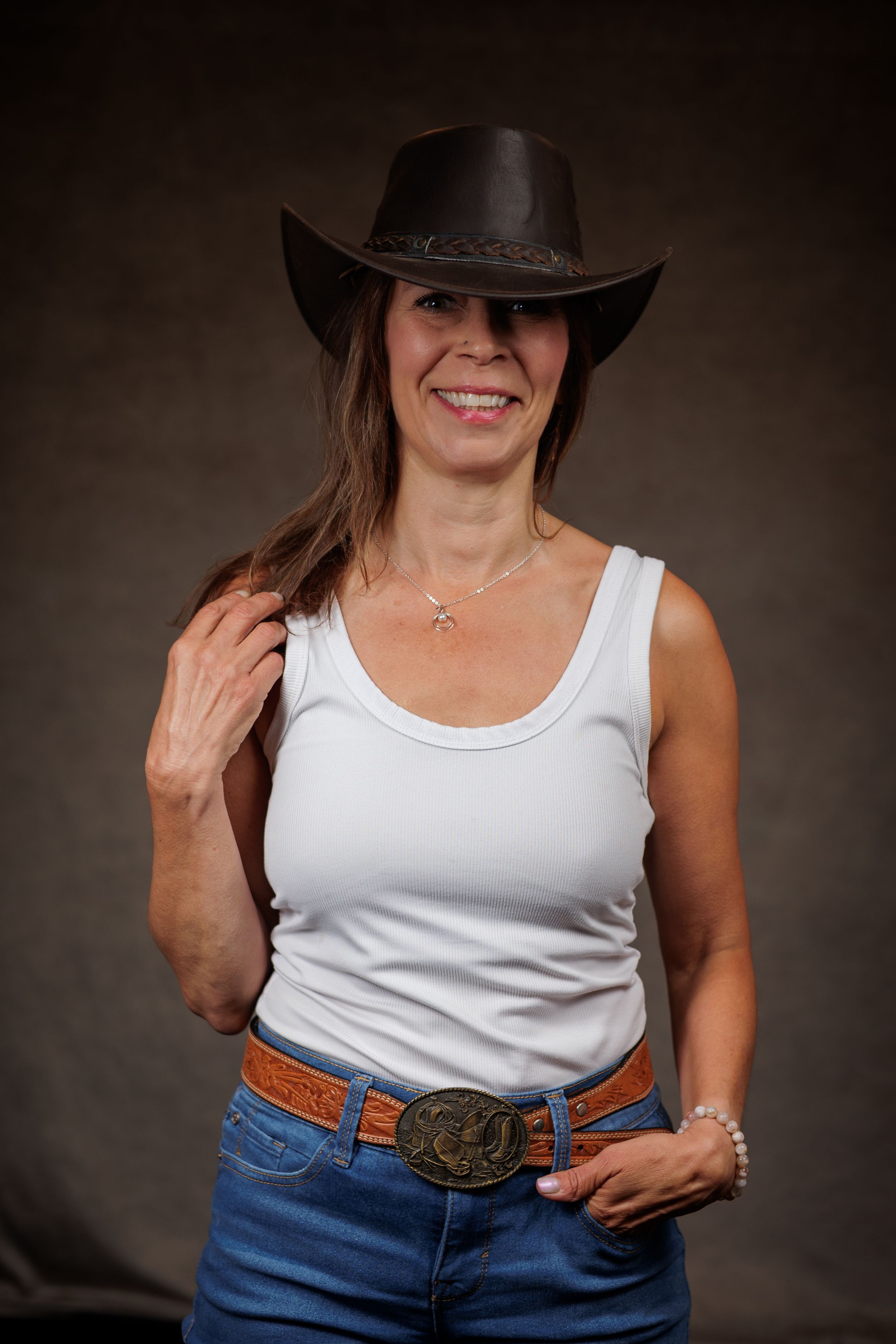A person wearing a cowboy hat, white tank top and jeans with a decorative belt buckle poses against dark backdrop.