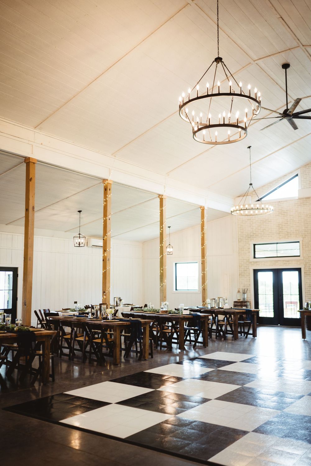 Elegant event venue with high ceilings, crystal chandeliers, checkered floors and rustic wooden posts features farm tables.