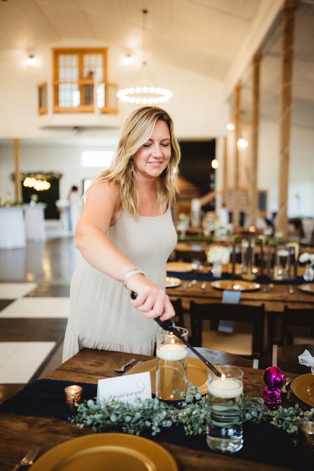 Person arranging candles and greenery on wooden table in rustic-style event venue with warm lighting.