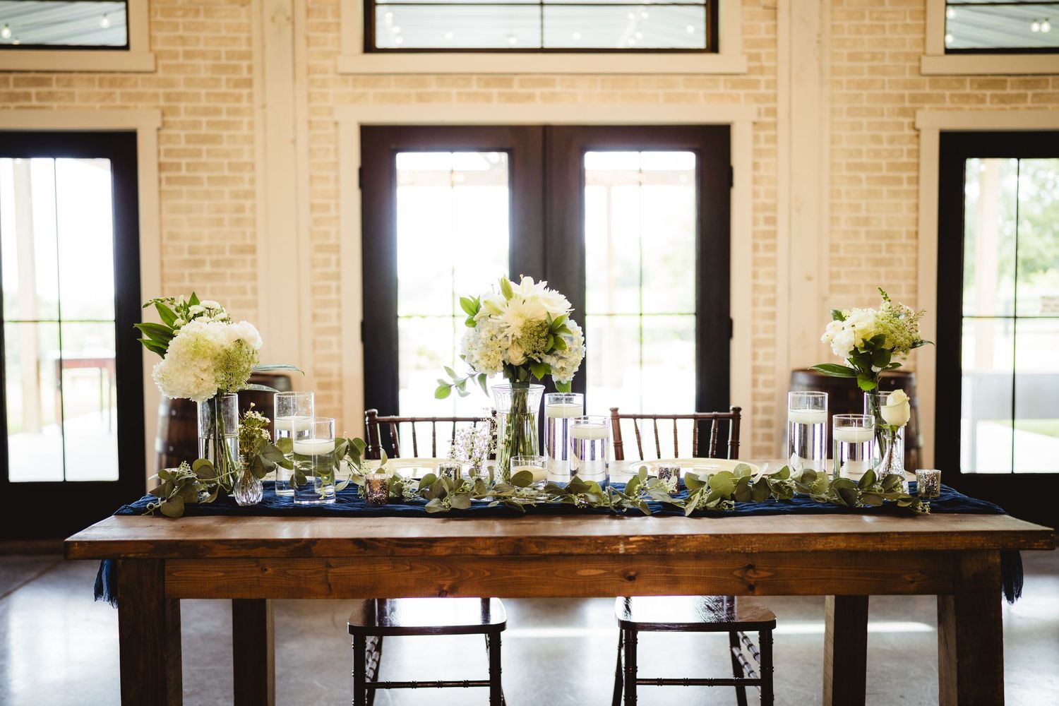 Dark wooden banquet tables in an indoor venue with floral arrangements and decorative settings, surrounded by natural light.