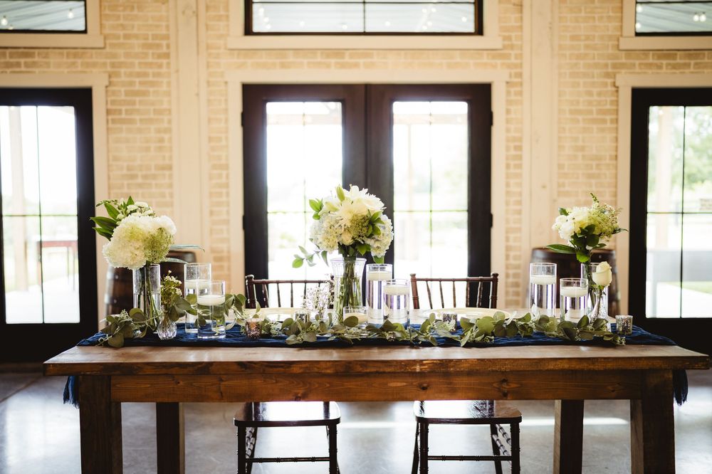 Dark wooden banquet tables in an indoor venue with floral arrangements and decorative settings, surrounded by natural light.