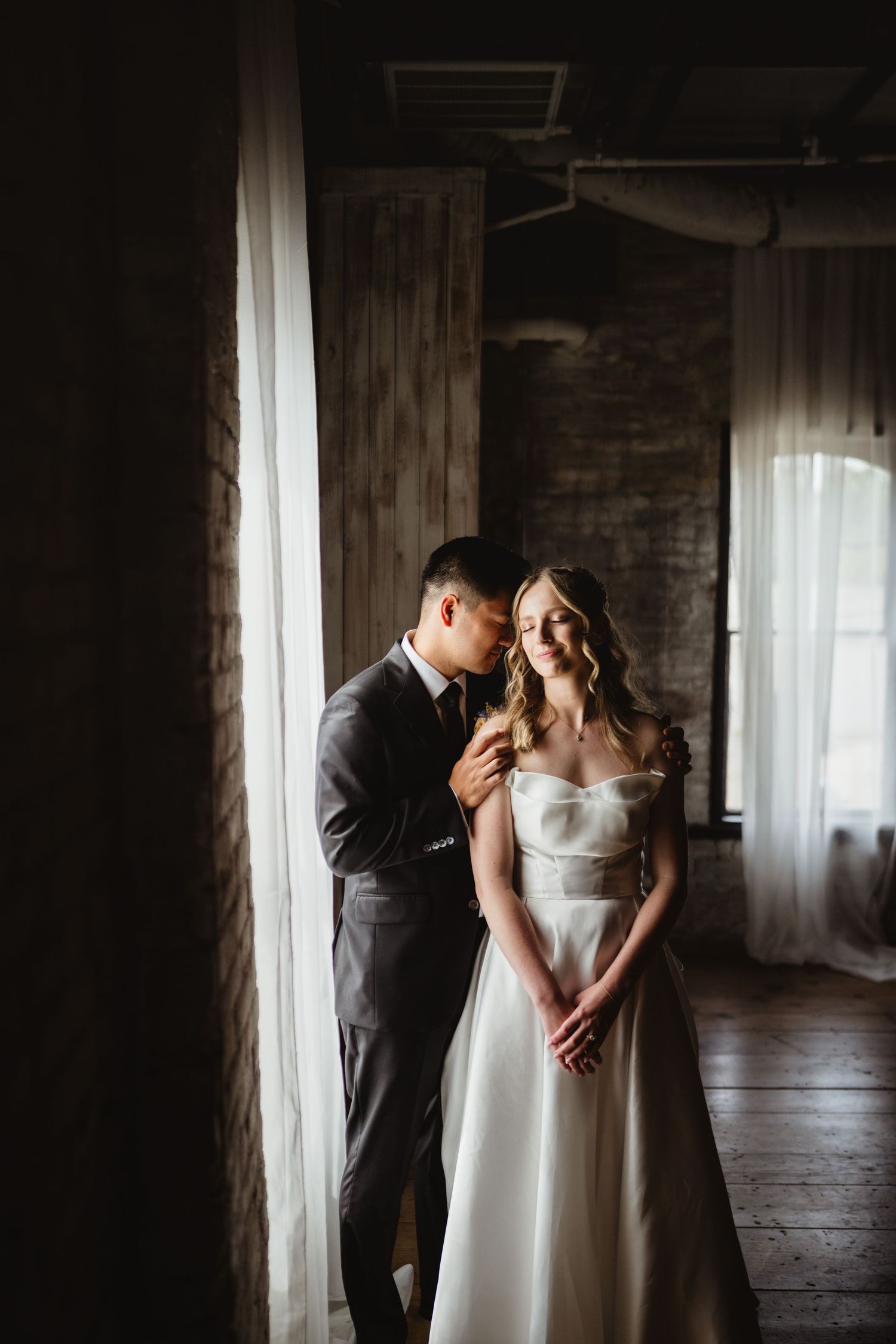 A couple shares intimate moments in a sunlit room with sheer curtains while dressed in formal wedding attire.